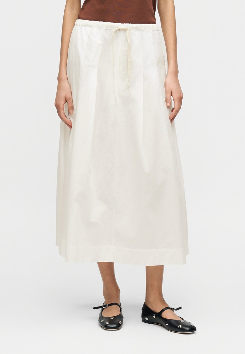 Λευκή φούστα μέχρι το γόνατο από ελαφρύ ύφασμα με ελαστική μέση και κορδόνι. Φορεμένη με μαύρα, επίπεδα παπούτσια Mary Jane με διακοσμητικά στοιχεία.