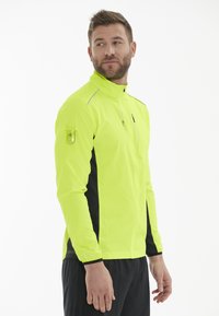 Herre træningsjakke i klar neon gul med sorte sidepaneler, med lynlås foran og en lynlåslomme på ærmet til opbevaring.