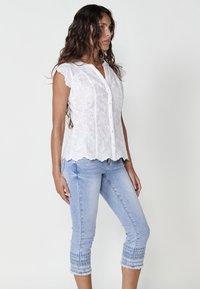 Blouse blanche brodée de fleurs avec des manches courtes, un ourlet festonné et une fermeture à boutons, associée à un jean capri bleu clair avec des poignets à motifs.