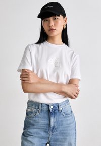 Hvit bomull t-skjorte med metallisk logo, kombinert med høydeydde lyseblå denimjeans. Svart caps og hoop-øredobber fullfører antrekket.