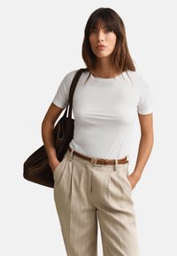 Femme aux cheveux bruns porte un haut blanc à manches courtes, un pantalon beige à fines rayures avec une ceinture marron, et porte un grand sac à épaule marron foncé.