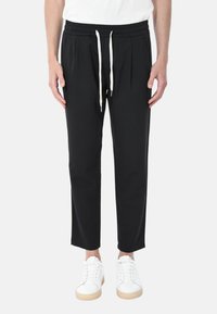 Pantaloni neri a sigaretta con vita elastica e coulisse, caratterizzati da pieghe sulla parte anteriore, abbinati a sneakers bianche.