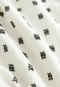 Vitt tyg med svarta rosettmotiv, med en lätt textur och ett subtilt mönster av broderade detaljer över ytan.