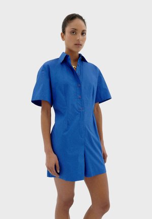 Combinaison bleue à manches courtes avec un col, fermeture à boutons sur le devant et une coupe ajustée. Tissu lisse avec un léger éclat.