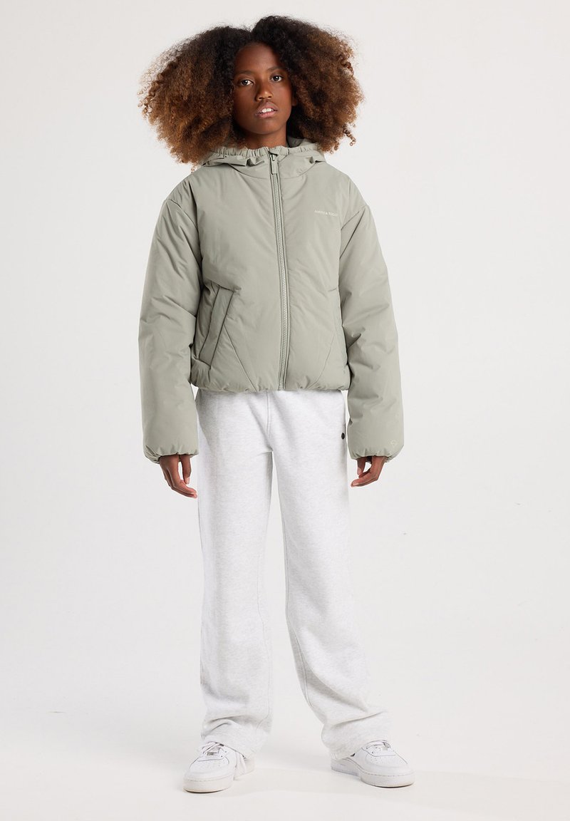Jong persoon met krullend haar, gekleed in een lichtgrijze capuchonjas, witte joggingbroek en witte sneakers, staand tegen een effen achtergrond.