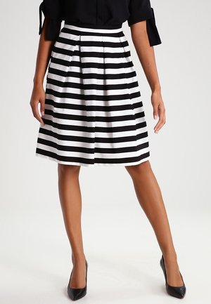 Femme portant une jupe rayée horizontale noire et blanche longueur genoux, un haut noir avec des manches à nœud, et des talons hauts noirs pointus.