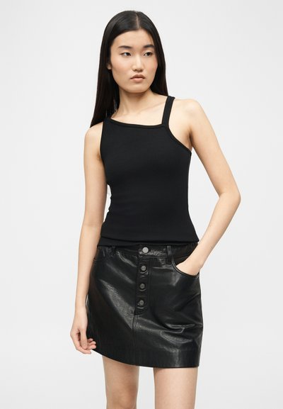 Jeune femme aux longs cheveux foncés portant un débardeur noir asymétrique à bretelle et une mini-jupe en cuir noire à boutons sur le devant, main dans la poche.