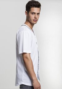 White short-sleeved button-up shirt with a V-neck, featuring black piping along the placket. Made from a smooth, lightweight fabric.