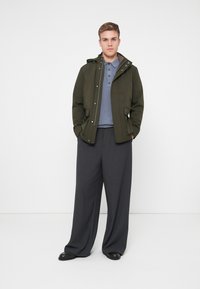 Olivovo zelená bunda s kapucňou, zapínanie na gombíky vpredu a bočné vrecká, oblečená cez modré polo tričko a široké sivé nohavice, v kombinácii s čiernymi topánkami.