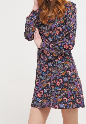 Femme portant une robe à manches longues, longueur genoux, avec un motif coloré de cachemire et fleurs sur fond noir.