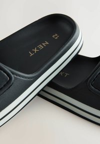 Sorte slip-on sko med en tekstureret overflade, der har hvide striber på sålen og guldskrift, der stavner "NEXT" indeni. Størrelse 12.