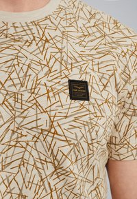 Beige kortærmet t-shirt med et kaotisk mønster af tynde, brune og grå linjer. Indeholder et lille sort mærke i stof på fronten.