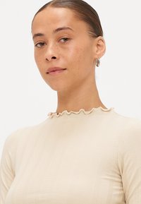 Beige båndet langærmet top med en volang-krave og glat tekstur, parret med sølvøreringe.
