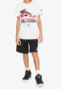 Camiseta blanca con un gráfico pixelado de unas zapatillas rojas y negras y el texto "AIR JORDAN". Pantalones cortos negros, zapatillas negras y calcetines blancos.