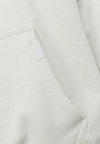 Lysegrå sweatshirt med kengurulomme, tekstureret stof og et lille vævet mærke på kanten. Blødt og afslappet design.