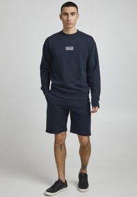 Sudadera azul marino con un gráfico blanco en el pecho, combinada con pantalones cortos azul marino. Cuenta con puños y dobladillo acanalados, llevada con zapatillas negras.