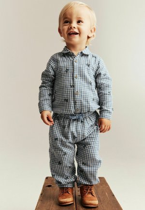 Kleuter met blond haar, gekleed in blauw geruite pyjama en bruine schoenen, staand op een houten oppervlak, glimlachend en omhoog kijkend.