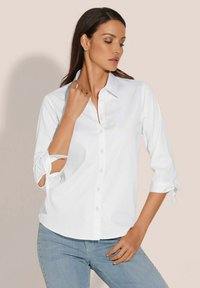 Witte blouse met een puntkraag, driekwart mouwen met strikjes en een gladde textuur, gecombineerd met lichtblauwe jeans.