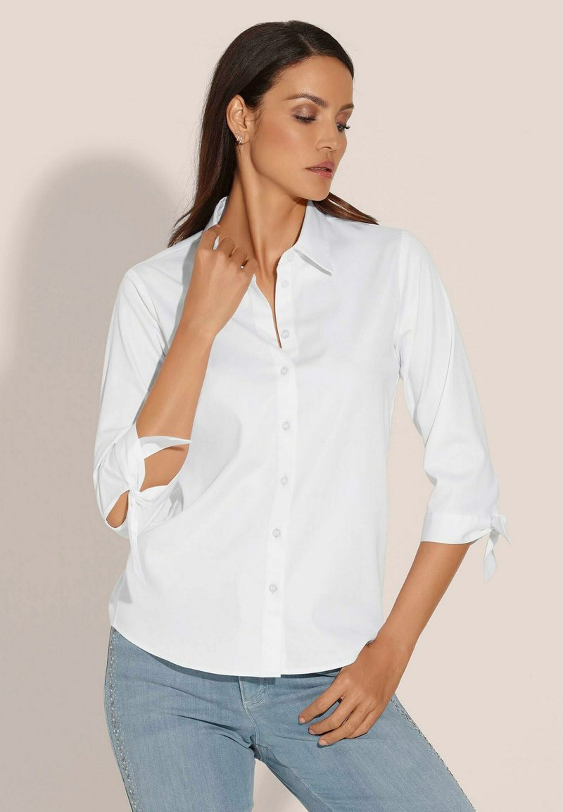 Witte blouse met een puntkraag, driekwart mouwen met strikjes en een gladde textuur, gecombineerd met lichtblauwe jeans.