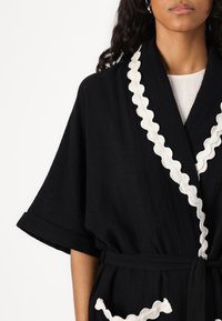 Robe noire à manches larges, avec un tissu texturé et un bord festonné blanc le long des bords et des poches. Nouée à la taille.