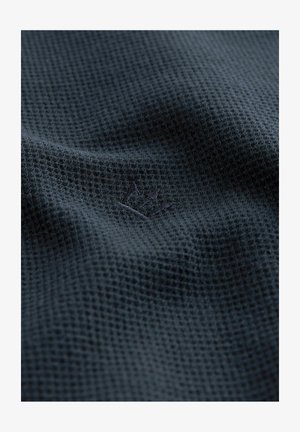 Tissu en tricot gaufré bleu marine avec une texture douce, arborant un logo brodé dans le coin. Le matériau est respirant et léger.