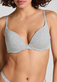 Eine Frau trägt einen hellgrauen gepolsterten BH mit dünnen Trägern, der den oberen Torso von Kinn bis Taille zeigt.