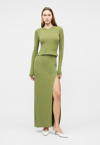 Γυναίκα που φοράει μακρυμάνικη μπλούζα σε ελιά πράσινο και μακρύ φόρεμα με σκίσιμο στο μηρό, συνδυασμένα με λευκά παπούτσια mule με τακούνι.