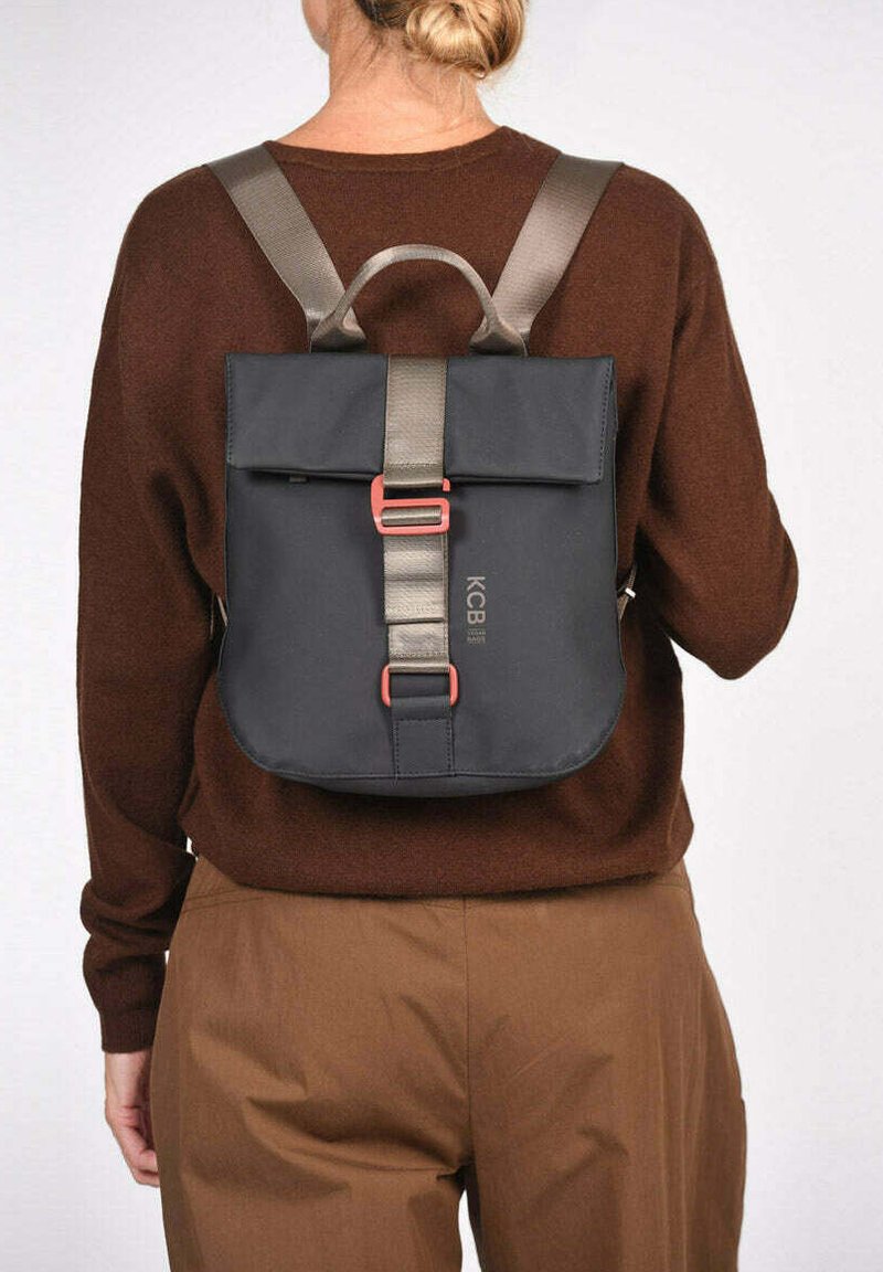 Mochila negra con solapa plegable y correas gris claro. Cuenta con una hebilla roja y detalle de logo. Textura suave, diseño compacto.