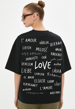 Donna con occhiali da sole e chignon che indossa una maglietta nera stampata con la parola "love" in più lingue sulla schiena.
