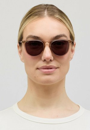 Frau mit blondem Haar, die runde, braun getönte Sonnenbrillen und ein weißes Oberteil trägt, nach vorne blickend vor einem einfarbigen hellen Hintergrund.