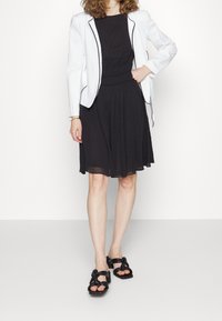 Černé šaty s plisovanou sukní, doplněné bílým blejzrem s černými paspulemi a černými sandály na vysokém podpatku s uzlovým designem.