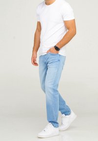 Man wearing a white t-shirt, light blue jeans, white sneakers, and a black smartwatch with one hand in pocket, standing on a light background.