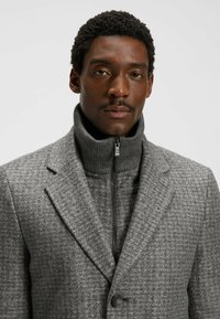 Man med kort, lockigt hår och mustasch, klädd i en grå mönstrad jacka över en grå zip-up polotröja.