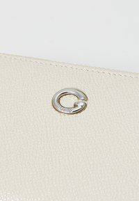 Portefeuille en cuir crème avec une surface texturée, doté d'un élément en métal argenté rond avec un logo gravé. Design compact et élégant.
