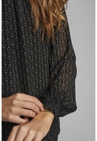 Blusa nera trasparente con motivo geometrico in oro e argento. Presenta polsini elasticizzati e maniche fluide. Mano che sistema un bracciale al polso.