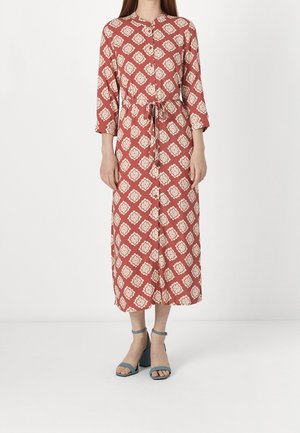 Kobieta w różowej sukience midi z kremowymi geometrycznymi wzorami, długimi rękawami, wiązaniem w talii oraz niebieskich sandałach na słupku z odkrytymi palcami.