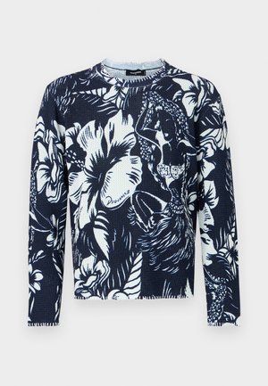 Ung mand med krøllet hår iført en marineblå og hvid blomstret sweater, slidte blå jeans og sorte læderstøvler, stående op ad en lys væg.
