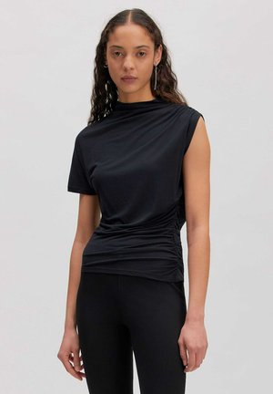 Femme aux longs cheveux bouclés portant un haut noir à manches courtes froncé et un pantalon noir sur un fond gris clair uni.