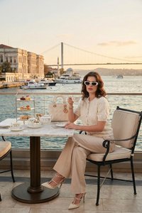 Femme en tenue beige et lunettes de soleil blanches sirotant un thé dans un café au bord de l’eau avec des pâtisseries, des bateaux et un pont suspendu en arrière-plan.