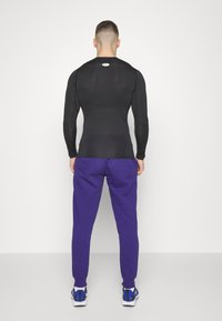 Svart långärmad träningsskjorta i kombination med lila träningsbyxor. Skjortan har en åtsittande passform med en mjuk textur och en logo på baksidan.