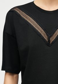 Μαύρο κοντομάνικο τοπ με ένθετο από δαντέλα σε σχήμα V στο ντεκολτέ που αποκαλύπτει το δέρμα από κάτω.