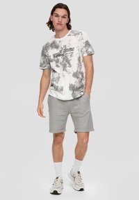 Szara i biała koszulka tie-dye z czarnym napisem, sparowana z jasnoszarymi szortami. Model ma na sobie białe skarpetki i sneakersy. Prosty, casualowy styl.