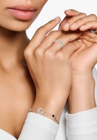 Gioielli in argento esposti su mani: un delicato bracciale a catena con un pendente ovale e un accento nero, e un anello rotondo con pietre preziose.