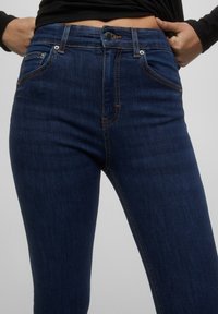 Jeans de mezclilla azul oscuro con cintura alta, que cuentan con un cierre de cremallera, botón de metal y costuras naranjas en contraste a lo largo de las costuras y bolsillos.