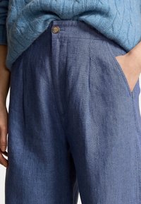 Modré lněné kalhoty mají volný střih, záhyby, zapínání na knoflík a boční kapsy, zachyceny vedle texturovaného modrého pleteného svetru.