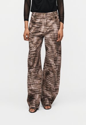 Παντελόνια με φαρδιά παντελόνα σε καφέ και μπεζ εκτύπωση ερπετού, συνδυασμένα με μαύρο διαφανές τοπ με μακριά μανίκια και μαύρα πέδιλα ανοιχτής μύτης.