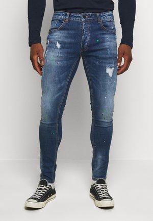 Slim fit blauwe jeans met versleten plekken en verfspatten, gedragen met zwarte sneakers en witte sokken door een staande persoon.