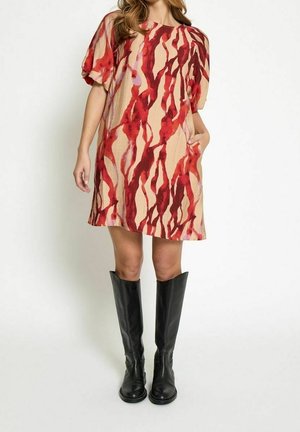 Femme portant une robe beige avec des motifs abstraits rouges, des manches courtes bouffantes, la main dans la poche et des bottes en cuir noires montantes jusqu’aux genoux.