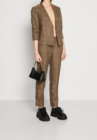 Brun rutig kavaj och byxset, kombinerat med en beige topp. Accessoarer inkluderar en liten svart handväska och robusta svarta skor.