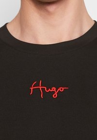 Svart bomullströja med en röd broderad logotyp "Hugo" på bröstet, rundad halsringning och mjuk textur.